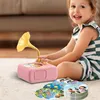 A young girl is playing with a pink toy gramophone and story cards on a carpeted floor. Produktbild-Vorschau
