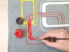 A hand is painting a red line onto a gray fabric with a pattern of yellow and white shapes. A small jar of red paint and its lid are also visible. Anteprima dell’immagine prodotto