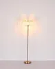 A floor lamp with a gold-colored metal base and stem, and a cylindrical glass shade with vertical ridges, is shown against a white wall. Náhľad obrázku produktu