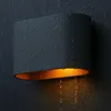 A dark, textured wall with rain falling, featuring a black and gold outdoor wall light with water droplets on its surface and water dripping from the bottom. Produktbild-Vorschau