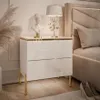 A white high-gloss nightstand with gold accents and legs, placed next to a bed in a bedroom setting. Produktbild-Vorschau