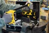 A person wearing gloves is using a yellow and black clamp tool to hold a metal piece in a vise. A toolbox with tools is in the background. Produktbild-Vorschau