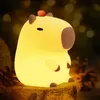 A glowing capybara-shaped night light sits on a wooden surface, with a blurred background of warm lights. Náhled obrázku produktu