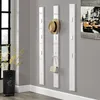 A hallway with white coat racks mounted on a gray wall. A straw hat hangs on one rack, and a beige purse hangs on another. A black planter with yellow flowers is on the left. Produktbild-Vorschau