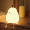 A pear-shaped LED night light with a smiling face sits on a wooden surface next to a yellow vase with greenery. Produktbild-Vorschau