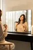 A woman is using a curling iron in front of a vanity mirror with lights. Produktbild-Vorschau