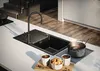 A modern kitchen scene featuring a black granite sink with a sleek faucet, two cups of coffee, and a bowl of cookies on a white countertop. A pot of liquid is on a drying rack next to the sink. Produktbild-Vorschau