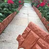 Braune Kunststoff-Gartenumrandung in Ziegeloptik, mit einem Stück im Vordergrund, installiert entlang eines gepflasterten Weges mit roten Rosensträuchern Náhľad obrázku produktu