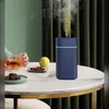 A person is holding a glass of tea next to a blue aroma diffuser on a marble table. There are also some chocolates and a lemon on the table. Produktbild-Vorschau