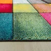 A close-up, angled view of a colorful, geometric patterned rug with a textured surface, placed on a light wooden floor. Produktbild-Vorschau