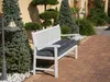 A white garden bench with a dark gray cushion sits on a brick patio surrounded by evergreen trees and a lamppost. Náhled obrázku produktu