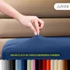 A hand is stretching a dark blue fitted sheet, with a row of different colored fitted sheets shown below. Produktbild-Vorschau