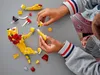 A child's hands assembling a yellow and red LEGO lion figure on a gray surface. Náhled obrázku produktu