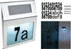 A collage showing a solar-powered house number sign, individual number stickers, mounting hardware, and a lifestyle shot of the sign mounted on a brick wall next to a blue door. Podgląd zdjęcia produktu
