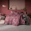 A bedroom scene with a bed dressed in red patterned bedding, a chair, and decorative items on a shelf. Produktbild-Vorschau