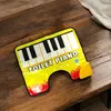 A yellow piano-shaped toilet mat with musical notes and flames is placed on a wooden table. Anteprima dell’immagine prodotto