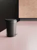 A black pedal bin is placed on a pink surface in front of a black and wood-paneled background. Voir les images du produit