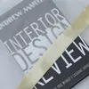 A close-up view of two books with a satin ribbon draped across them. One book has the title 'INTERIOR DESIGN' and the other has 'REVIEW' visible. Produktbild-Vorschau