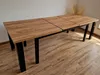 A long, extendable dining table with a wood-grain top and black metal legs, set against a plain wall and wooden floor. Produktbild-Vorschau