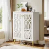 A white cabinet with decorative doors and gold legs, placed in a room with curtains and a window. Produktbild-Vorschau