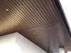 A view of a brown slatted ceiling or soffit, likely made of plastic or composite material, with integrated lighting. A white wall and a downspout are also visible. Produktbild-Vorschau