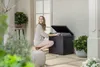 A woman is crouching and putting items into a black storage box on a patio. Podgląd zdjęcia produktu