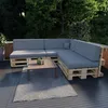 An outdoor seating area with a sectional sofa made of pallets and grey cushions, a coffee table, and a rug on a dark deck, surrounded by lush greenery. Produktbild-Vorschau