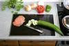 A kitchen counter with a black induction hob, a cutting board with sliced cucumber, smoked salmon, and a knife. In the background are onions, red and green bell peppers, and a potted plant. Anteprima dell’immagine prodotto