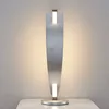 A modern LED table lamp with a silver finish is displayed on a white surface against a textured wall. Produktbild-Vorschau