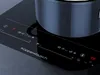 A close-up of a modern black induction cooktop with a pot on it, showing the control panel and brand name. Produktbild-Vorschau