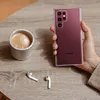 A hand holding a transparent phone case with a burgundy phone inside, next to a cup of coffee and wireless earbuds on a wooden table. Produktbild-Vorschau