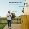 A man is standing on a wooden deck, holding a tablet and looking at a security camera mounted on a wooden house. The camera has a solar panel. Speech bubbles indicate a two-way audio conversation. Produktbild-Vorschau
