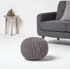 A knitted pouf is placed on a white floor in front of a gray sofa and a small wooden side table with a potted plant. Náhled obrázku produktu