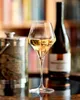 A glass of white wine is in the foreground, with a bottle of wine and an ice bucket in the background. The background is blurred, showing shelves with books or decorative items. Produktbild-Vorschau