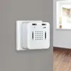 A white emergency alert device is mounted on a gray wall, with a window and wooden floor visible in the blurred background. Produktbild-Vorschau