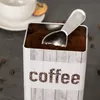 A coffee scoop is inside a coffee container, with a cup and cookie in the background. Produktbild-Vorschau