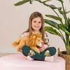 A young girl sits cross-legged on a pink surface, holding a stuffed lion. A large plant is to her right. Voir les images du produit