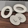 Two white ear pads and two gray fabric inserts with L and R markings are placed on a wooden surface in front of a laptop keyboard. Anteprima dell’immagine prodotto