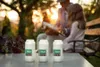 Three white candles are in the foreground on a table, with a blurred background of people sitting on a bench in a garden during sunset. Podgląd zdjęcia produktu