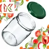 A glass jar and its lid are shown, surrounded by various fresh vegetables and herbs. Podgląd zdjęcia produktu