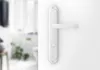 A close-up of a white door handle on a white door, with a blurred background of a room. Produktbild-Vorschau