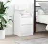A white bedside table with two drawers and a small shelf on top, next to a bed. A large potted plant is on the left. Náhľad obrázku produktu