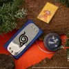 Naruto-themed merchandise, including a headband, a patch, and a tape measure, arranged on a textured surface with greenery. Anteprima dell’immagine prodotto
