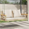 A beige poly rattan hanging bench with white cushions is shown on a wooden deck, with a wooden fence and greenery in the background. Náhled obrázku produktu