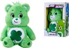 A green Care Bear plush toy with a shamrock on its belly, shown next to its packaging. Produktbild-Vorschau