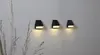 Three black solar wall lights mounted on a textured white wall, with dried grass in the foreground. Produktbild-Vorschau