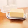 A block of butter sits in a clear plastic butter dish with a clear lid, placed on a wooden surface. Produktbild-Vorschau