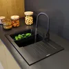 A modern kitchen sink with a black faucet running water into a black sink basin. Cucumbers are in a black colander within the sink. Jars of nuts and wooden bowls are on the counter behind the sink. Podgląd zdjęcia produktu