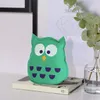 A green owl-shaped air freshener sits on a book next to a vase with eucalyptus leaves. Produktbild-Vorschau