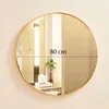 A round gold-framed mirror is shown hanging on a wall, reflecting a bathroom interior with a plant and tissue box. Produktbild-Vorschau